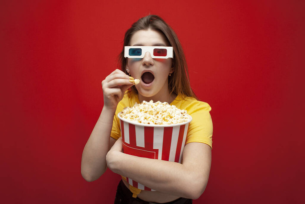 beautiful young emotional girl in 3D glasses eats popcorn, watches a movie and screams on a red color background, surprised viewer - Foto, Imagem
