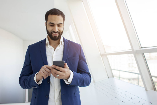 ελκυστική επιτυχημένη επιχειρηματίας σε ένα κοστούμι χρησιμοποιούν ένα smartphone σε ένα λευκό γραφείο και εκτυπώνει ένα μήνυμα - Φωτογραφία, εικόνα