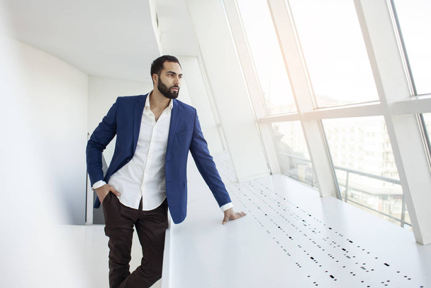 όμορφος επιτυχημένος διευθυντής εταιρείας στέκεται σε ένα λευκό γραφείο και κοιτάζει έξω από το παράθυρο, πορτρέτο ενός επιχειρηματία στο χώρο εργασίας - Φωτογραφία, εικόνα