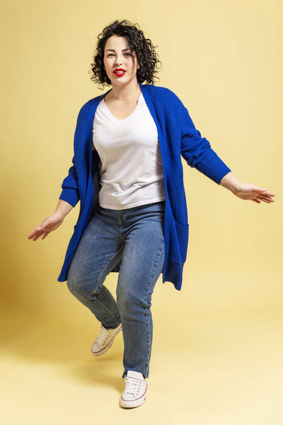 Dancing and laughing beautiful young woman plus size in jeans and a blue sweater. Yellow background. - Foto, Imagem