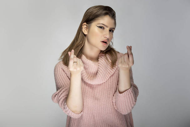 Close up portrait of a young Caucasian woman wearing a pink sweater on a grey background.  Her expressions is a money gesture - Photo, Image