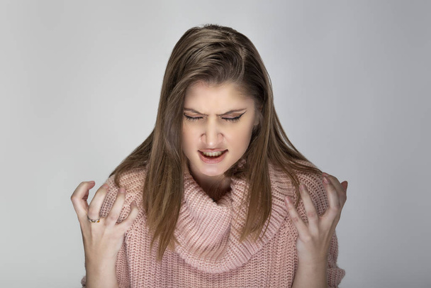 Close up portrait of a young Caucasian woman wearing a pink sweater on a grey background. She looks angry and in stress - Foto, imagen
