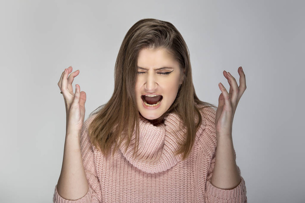 Close up portrait of a young Caucasian woman wearing a pink sweater on a grey background. She looks angry and in stress - Foto, imagen