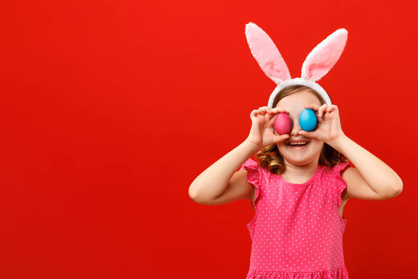 Happy cheerful little girl holding two easter eggs. Close up portrait of a child on a red background. Copy space - Foto, afbeelding