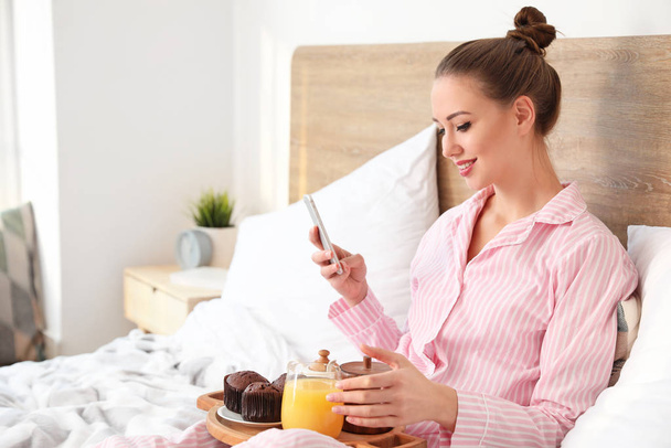 Poranek pięknej młodej kobiety z telefonem komórkowym jedzącej śniadanie w łóżku - Zdjęcie, obraz