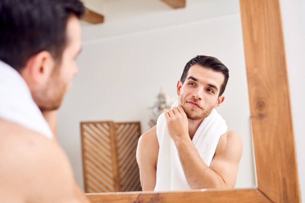 Unshaven undressed man with white towel on his neck standing near mirror - Foto, afbeelding