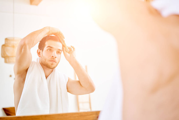 Young man combing his hair standing near mirror in bathroom in morning - Fotografie, Obrázek
