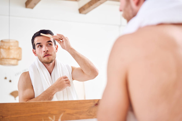 Young unshaven man combing his hair while standing with white towel on neck near mirror in bathroom - Foto, Imagem