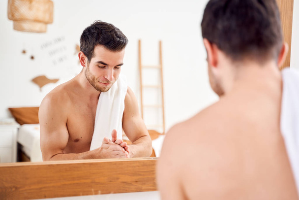 Young sports man lathers hands and stands with towel on his shoulders in front of bathroom mirror - Фото, изображение