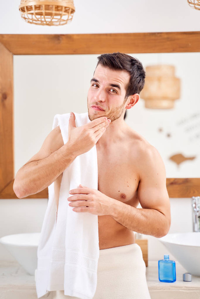 Male washes his face and stands with towel on his shoulders in front of bathroom mirror - Foto, immagini