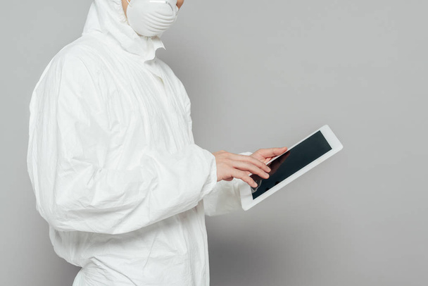 vista recortada del epidemiólogo en traje de materiales peligrosos y máscara respiratoria que sostiene la tableta digital con pantalla en blanco sobre fondo gris
 - Foto, Imagen