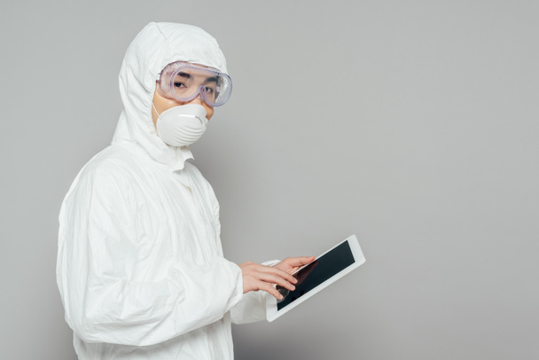asian epidemiologist in hazmat suit and respirator mask holding digital tablet with blank screen and looking at camera on grey background - Photo, Image