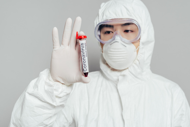 ázsiai epidemiológus védőruhában és légzőmaszkban vérmintával ellátott kémcsőt mutat szürke alapon - Fotó, kép
