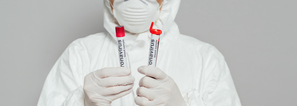 oříznutý pohled na epidemiologa v ochranném obleku a respirační masce držícího zkumavky se vzorky krve izolovanými na šedém, panoramatickém záběru - Fotografie, Obrázek