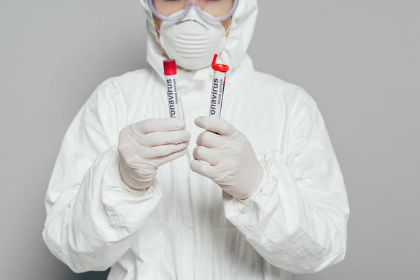 cropped view of epidemiologist in hazmat suit and respirator mask holding test tubes with blood samples on grey background - Photo, Image