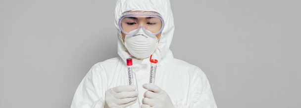 ハズマットスーツのアジア疫学者とマスクのパノラマ写真灰色で隔離された血液サンプルの検査管を保持 - 写真・画像