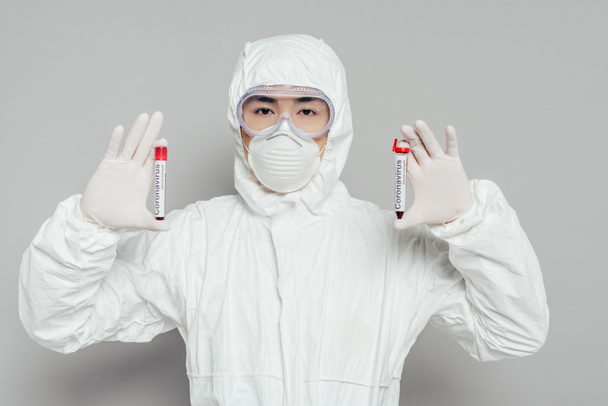 epidemiologo asiatico in tuta hazmat e maschera respiratoria che mostra provette con campioni di sangue su fondo grigio
 - Foto, immagini