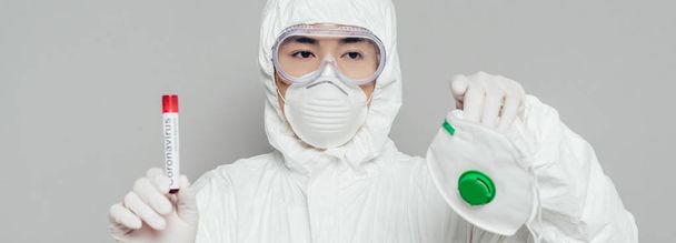 tiro panorâmico de asiático epidemiologista mostrando respirador máscara e tubo de ensaio com amostra de sangue isolado em cinza
 - Foto, Imagem