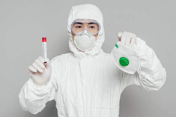 asian epidemiologist in hazmat suit holding respirator mask and test tube with blood sample on grey background - Photo, Image