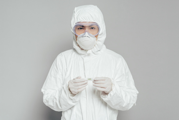 asiático epidemiologista olhando para câmera enquanto segurando termômetro mostrando alta temperatura no fundo cinza
 - Foto, Imagem