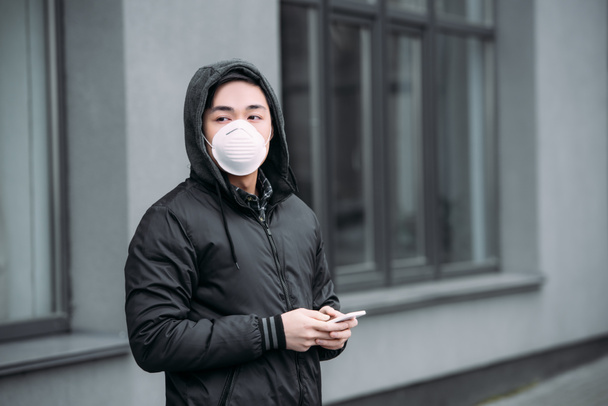 молодий азіатський чоловік в респіраторній масці тримає смартфон і дивиться геть, стоячи на вулиці
 - Фото, зображення