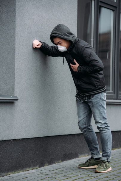 молодий чоловік в респіраторній масці спирається на стіну і торкається грудей, страждаючи від болю в легенях
 - Фото, зображення