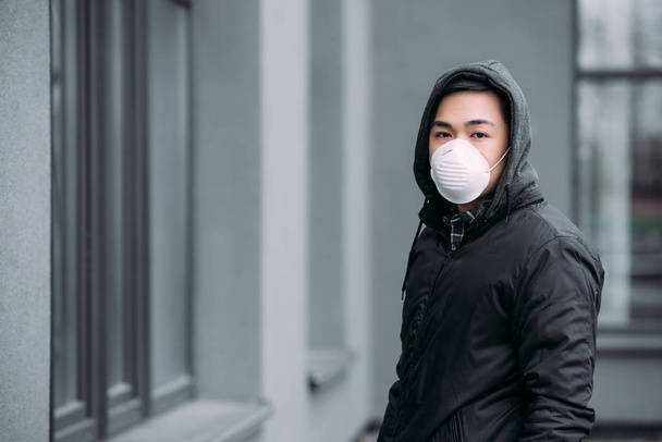 jong aziatische man in respirator masker kijken naar camera terwijl staan op straat - Foto, afbeelding