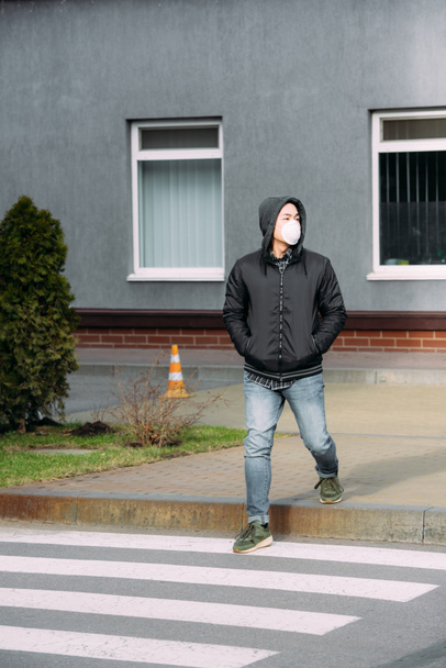joven asiático hombre en respirador máscara mirando lejos y cogido de la mano en bolsillos mientras cruzando camino
  - Foto, Imagen
