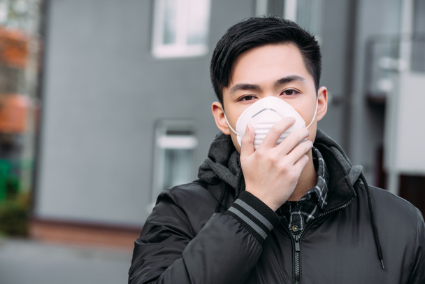 joven asiático hombre tocando respirador máscara y mirando a cámara mientras de pie en calle
 - Foto, Imagen