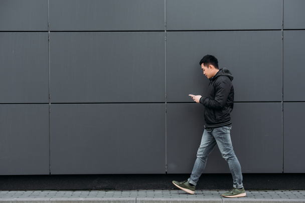 side view of young asian man chatting on smartphone while walking along wall - Photo, Image