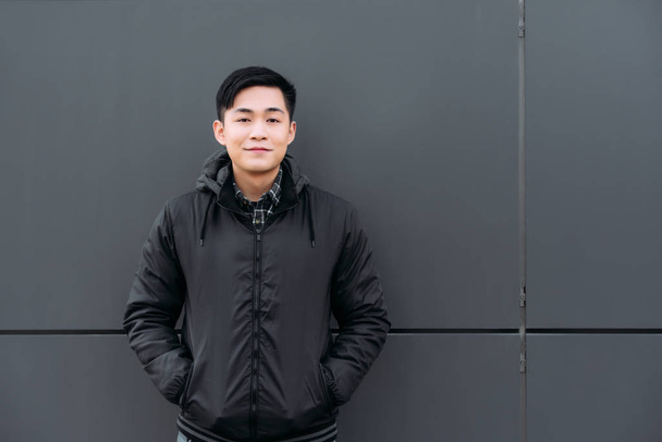 giovane asiatico uomo sorridente a macchina fotografica mentre in piedi da parete e tenendo le mani in tasche
 - Foto, immagini