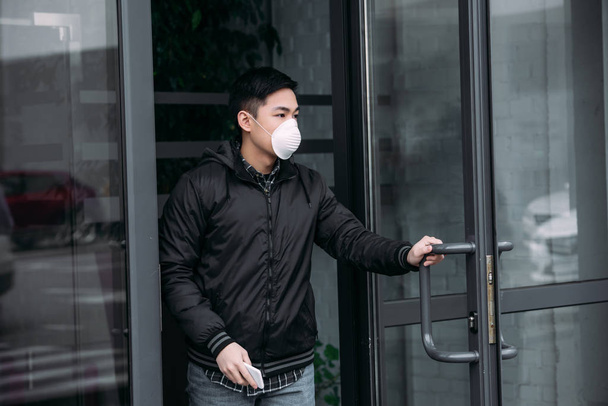 young asian man in respirator mask holding smartphone and looking away while leaving building  - Fotoğraf, Görsel