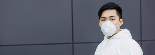 panoramic shot of asian epidemiologist in hazmat suit and respirator mask looking at camera while standing near wall - 写真・画像