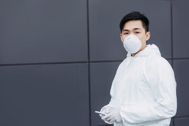 ázsiai epidemiológus vegyvédelmi ruha és légzőkészülék maszk gazdaság smartphone és nézi a kamera, miközben áll a fal közelében - Fotó, kép