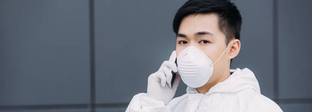 Panoramaaufnahme eines asiatischen Epidemiologen in Warnanzug und Atemschutzmaske, der mit dem Smartphone spricht und in die Kamera blickt - Foto, Bild