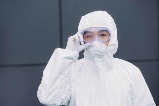 asian epidemiologist in hazmat suit and respirator mask looking at camera while standing outside and touching goggles - 写真・画像