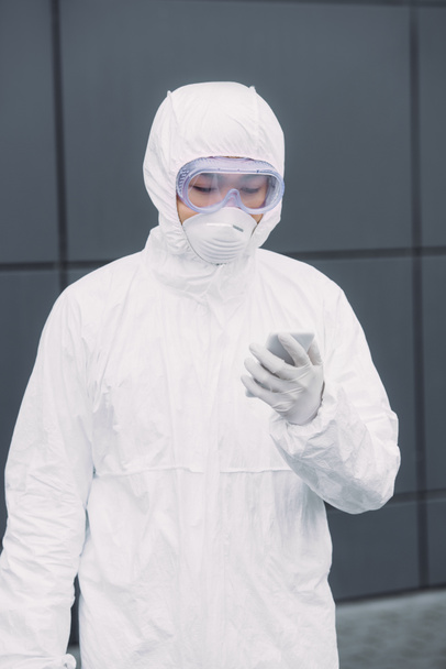 asian epidemiologist in hazmat suit and respirator mask standing outside and looking at smartphone - Фото, изображение