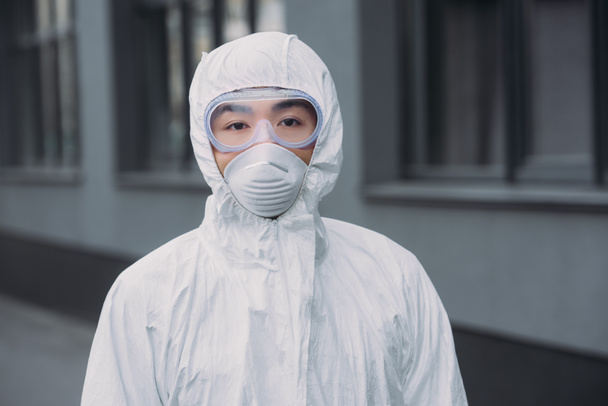 asiático epidemiólogo en hazmat traje y respirador máscara mirando a cámara mientras de pie cerca de la construcción
 - Foto, Imagen