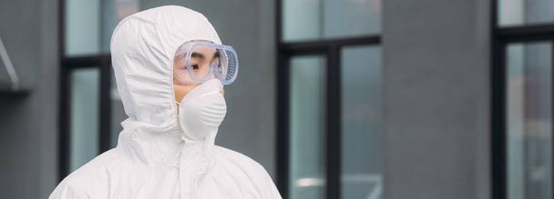 panoramic shot of asian epidemiologist in hazmat suit and respirator mask looking away while standing on street near building - Photo, Image