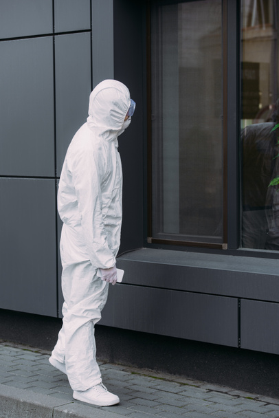 epidemiologist in hazmat suit standing on street and looking in window of building - Photo, Image