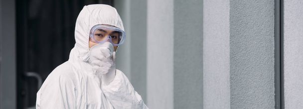 plano panorámico de epidemiólogo asiático en traje de hazmat y máscara respiratoria mirando a la cámara mientras está de pie fuera
 - Foto, Imagen