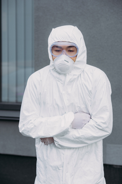 azjatycki epidemiolog w kombinezonie ochronnym i masce respiratora stojący ze skrzyżowanymi ramionami cierpiący na objawowy ból brzucha na ulicy - Zdjęcie, obraz