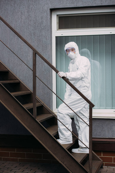 Asiatischer Epidemiologe in Warnanzug und Atemschutzmaske, der vor dem Gebäude die Treppe hochgeht - Foto, Bild