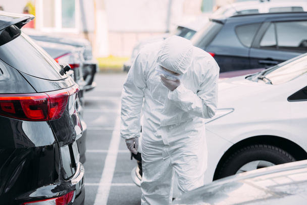 aziatische epidemioloog in hazmat pak en masker van de respirator inspecteren voertuigen op de parkeerplaats - Foto, afbeelding