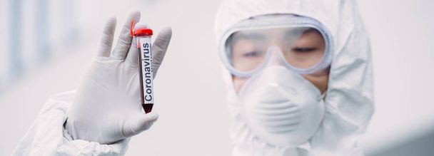 inyección panorámica de epidemiólogo asiático en traje de hazmat y máscara respiratoria mirando el tubo de ensayo con muestra de sangre mientras está de pie al aire libre
 - Foto, Imagen