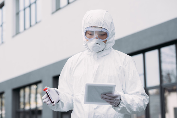 asian epidemiologist in hazmat suit and respirator mask using digital tablet while holding test tube with blood sample on street - Fotografie, Obrázek