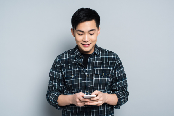 joven asiático hombre sonriendo mientras usando smartphone en gris fondo
 - Foto, Imagen