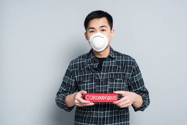 young asian man in respirator mask showing warning card with coronavirus inscription on grey background - Photo, Image