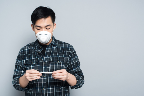 νεαρός ασιάτης με μάσκα αναπνοής που κοιτάζει το θερμόμετρο σε γκρι φόντο - Φωτογραφία, εικόνα