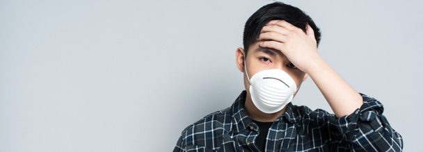 Panoramaaufnahme eines jungen asiatischen Mannes mit Atemschutzmaske, der die Stirn berührt und in die Kamera schaut, während er an Kopfschmerzen leidet, isoliert auf grau - Foto, Bild